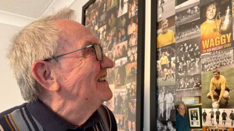 A bespectacled man in his 80s smiles as he focuses on a compilation of old photographs on the wall of his living room.
