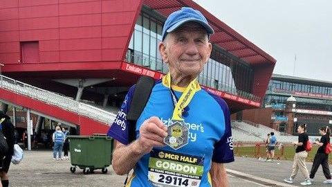 Gordon Aaron smiles and holds up his medal as he poses for a picture in Old Trafford cricket ground. He wears a blue cap and a blue top with Alzheimer's Society branded on it.