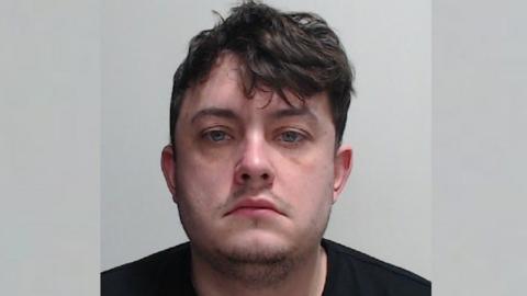 a police mugshot of a man with brown hair wearing a black t-shirt