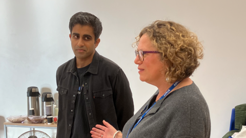 A man with dark hair, a dark shirt and a dark jacket in a white room with coffee urns behind him, looking at a woman with blond wavy hair and wearing a grey top