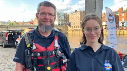 A man with grey hair and a beard looks into the camera. He is wearing a life jacket with equipment for saving lives. He is standing next to his 16-year-old daughter who is wearing a dark blue polo shirt with the "York Rescue Boat" logo on it.