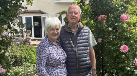 An older couple, one female and one male, David and Liz. They stand in front of an archway made of pink rose bushes. Liz has cropped grey hair and wears a floral blouse with glasses on a chain around her neck. David wears a checked shirt and dark body warmer. He wears clear glasses and has fine grey hair. Both smile.