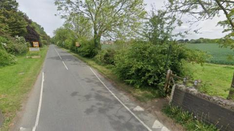 A Google street view image of Belvoir Road in Bottesford with a gate to a public foothpath on the right of the image and a sign saying "Bottesford" on the left, separated by a two-lane road. 