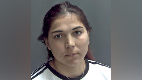 A custody photo of Cristina Curt. She has dark hair dyed slightly red and pulled into a pony tail and she is wearing a white t-shirt with black stripes