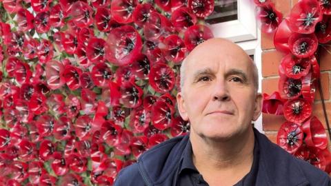 Chris Penrose is wearing a blue jacket and t-shirt. The red poppy display is behind him.