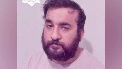 A man with dark hair and a full beard, wearing a light-coloured top against a plain background. A police crest is partially visible in the top corner of the image.