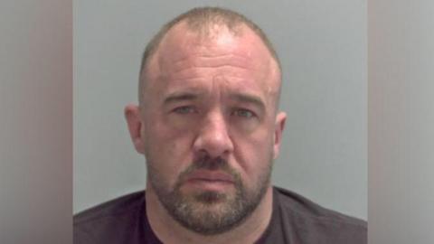 James Morgan has a short buzz cut and beard in his custody photo. He is wearing a brown T-shirt. 