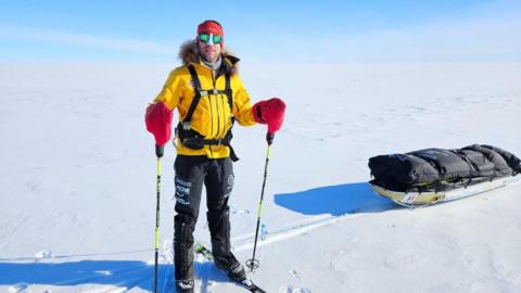 Jonny Huntington pictured on the ice wearing a bright yellow coat and black trousers. He is also seen wearing red gloves and a red hat with sunglasses.