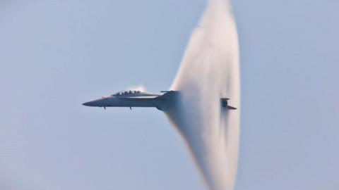 A grey fighter jet is flying through a large cloud-like pressure wave. There are blue skies behind.