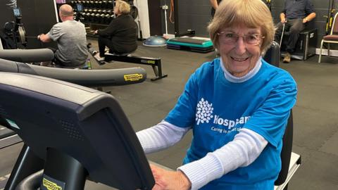 Dorrie sits on an exercise bike in a busy gym. She has dark blonde hair and glasses. She is wearing a blue Hospicecare t-shirt over a thin jumper. She is smiling at the camera.