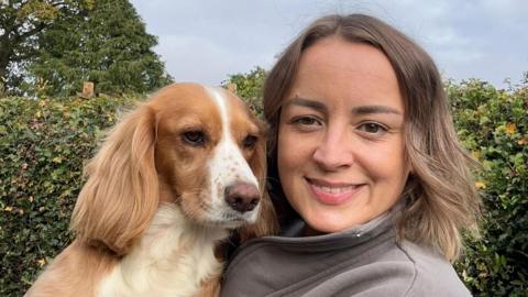 A smiling woman with shoulder length brown hair looks to camera. She is holding up a ginger coloured spaniel dog alongside her. She is stood in front of a green hedge.