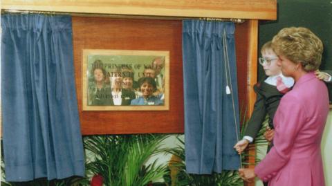 A woman with short blonde hair, wearing a rose-coloured jacket, holds a small child who has green-rimmed glasses and wears a suit-like jacket over a white shirt and pink tie. Both of them are pulling on a cord which has opened a pair of blue curtains to reveal a plaque