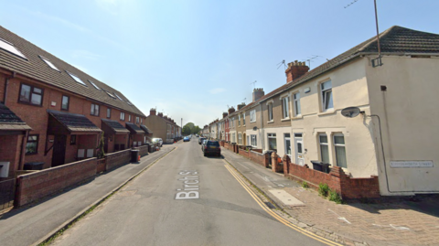 A screenshot of Google Streetview, showing Birch Road from one end. The picture was taken on a sunny day and shows the long, straight road. One the left there is a line of darker-coloured terraced houses, with lighter, older-looking terraces on the right. There are some cars parked along the road further down the street. 