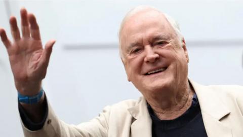 Actor John Cleese is seen smiling and waving at the camera. He has white hair and is wearning a cream suit jacket, blue watch and black jumper.