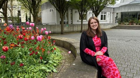 Jen is sitting on a concrete circle that surrounds a flowerbed. In the backdrop is a grey building. She is wearing a black academic gown and pink leopard print dress.