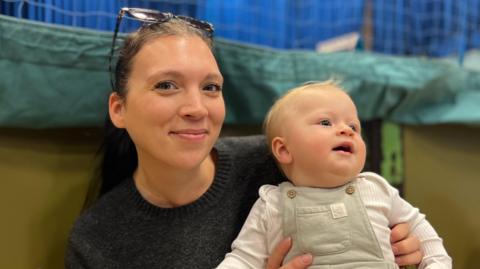 Leigh Walker and her baby Clyde - A woman with sunglasses on her head smiling at the camera. She's black hair in a ponytail and a black jumper. She is holding on her lap her baby who is wearing green dungarees and a white baby-gro. He's looking away from the camera and has a tuft of blonde hair on his wee head.