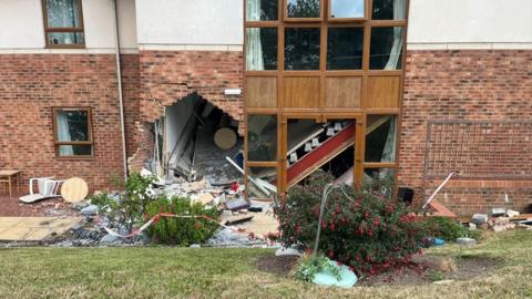 A large hole at the bottom left of a brick and white stone building next to a window. Bricks and debris from the wall are scattered all around and in the bushes and grass in front of the building.