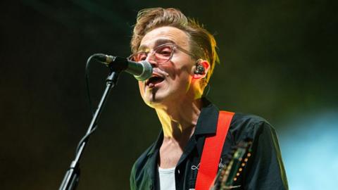 om Fletcher of McFly performs during Download Festival Donington Park on June 13, 2025 in Castle Donington, England. He has a black jacket on and is singing into a microphone while playing an electric guitar (which is just out of shot). You can see the red strap from his guitar though.