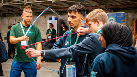 A boy aims a bow and arrow. Two other teenagers, a girl and a boy, look on standing either side of him. A man in the background wears green, appearing to be an instructor, and seems to be talking to them.