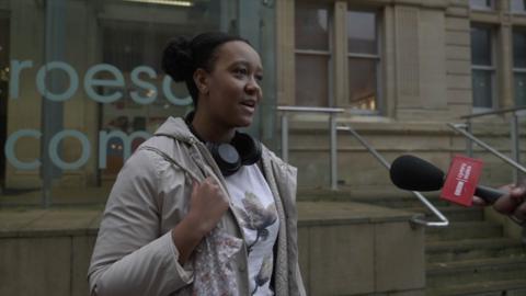 A young woman wearing a cream jacket and headphones. She is hold a floral bag around her shoulder