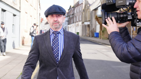 Terrence McBrien walking past walking past a cameraman in Berwick in March 2026. He is wearing a tweed cap, jacket, a striped blue shirt and a tie. He has blue eyes and thin greying beard.
