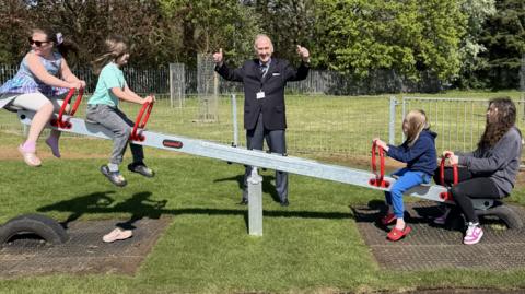 Four children on sea saw, with a man in the middle, with his arms up, wearing a black suit. The park has lots of green grass and trees. 