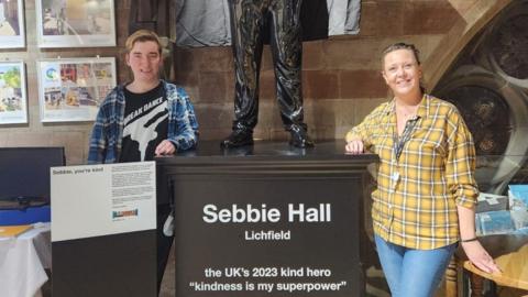 A man in a black tshirt with a blue checked shirt over the top of it stands next to a statue with a woman on the other side of the statue wearing a yellow checked shirt