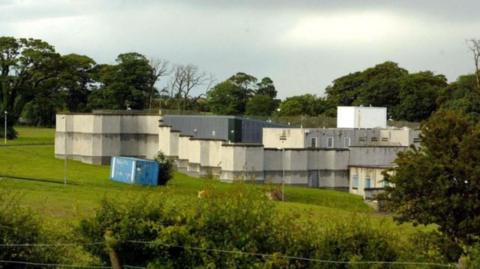 Landscape shot of Kerelaw residential school in Stevenston, North Ayrshire. The school is grey and white with a flat roof and is pictured behind a wire fence and hedge row, with a line of trees in the background.