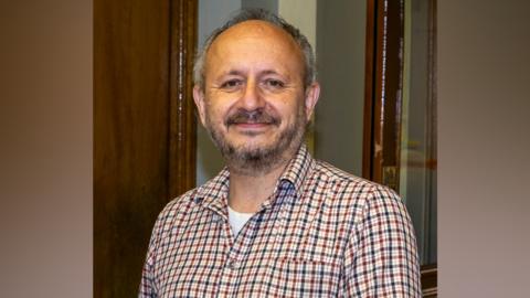 A man with grey hair and a grey beard, wearing a red and blue check shirt.
