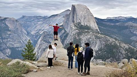People take photos from Glacier Point with Half Dome in the background at Yosemite National Park. A man in a red shirt is standing at the edge of a popular photo spot with is arms spread out.