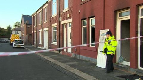 A police officer stands at the doorway of the fire-hit property