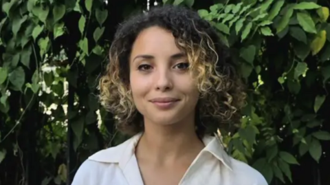 A woman standing against a backdrop of vibrant green foliage. They're wearing a white-coloured collared shirt, which creates a striking contrast with the lush greenery behind them. Her hair is curly and a blend of dark and lighter shades, adding to the texture of the scene.