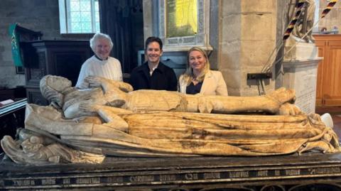 Two men and one woman standing in a church behind a stone tomb with the figures of a praying man and woman lying on top. One man has white hair and a white jumper, the other has dark hair and a dark jumper and the woman has blond hair and a white jacket