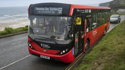 A red single-decker bus going along a costal road with a beach behind. The bus is the number 6 going to Newquay Bus Station via Constantine Bay,