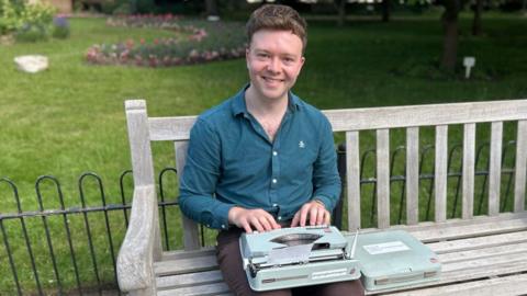 A man in brown trousers and navy shirt sits on a wooden bench, an old typewriter on his knee. Behind him are low iron railings, grass, and a flower bed.