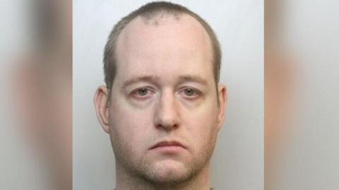 Mugshot of a man wearing a grey shirt. He has very short hair and stubble. 