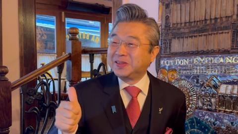 The Japanese ambassador smiles while holding his thumbs up in a cafe in Liverpool