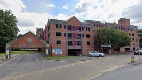 A street view image of a brick-built multi-storey car park. On the left is an entrance with a brick sign overhead that says "St Martin's Gate". Two cars can be seen coming out of the car park's exit on the right.