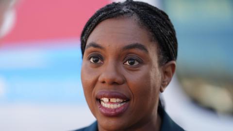 Kemi Badenoch is speaking into a camera and is looking slightly off to the left of the frame. She is wearing a blue jacket which can be slightly out of shot.