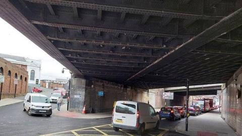 The underneath of a bridge with large steel beams above it. There are cars queueing in a line underneath it, as well as a van waiting on the left