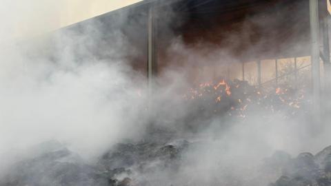 A pile of straw which was sat inside and outside of a barn was on fire. Grey smoke was coming from the piles.