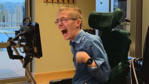 A man sitting down in a green wheelchair, with glasses wearing a blue shirt with white detailing. He is very smiling and visibly happy.