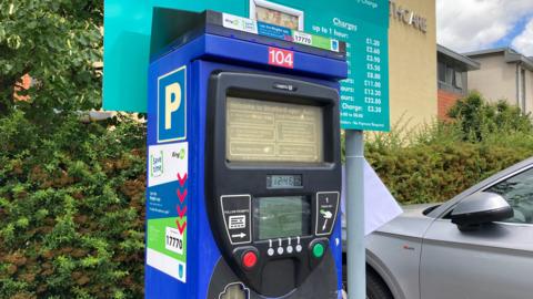 A blue car park park ticket machines with black inset and green and red push buttons, behind it is a large turquoise sign displaying car park fees.
