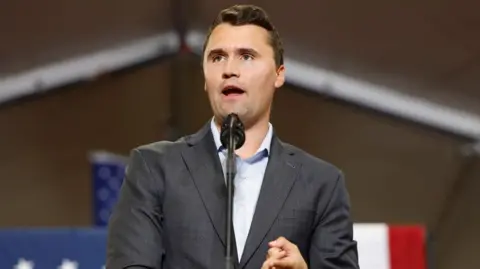 Charlie Kirk, wearing a grey suit, speaks at a podium