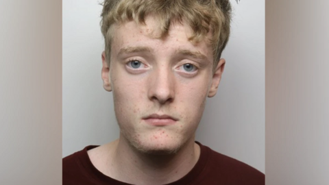 A police mugshot of a man with short blonde hair and a dark red t-shirt.