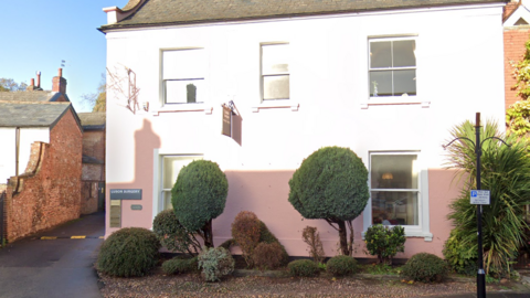 A screenshot of Google street view. It's the external of the building, which is painted pink, and the sun is making the top half of the building very bright. 