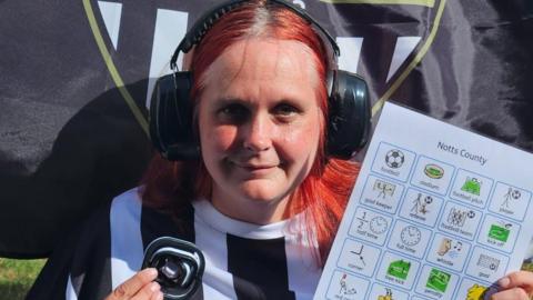 Laura Cameron is pictured wearing headphones and a Notts County home shirt. She has red hair and also holds an A4 laminated card with different symbols.