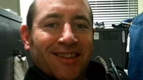 A selfie taken by David Carrick of him sitting in an office with a filing cabinet behind him. He is smiling and has a walkie-talkie strapped to his chest.