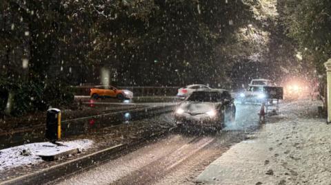 Several cars driving through snow in Leamington, there is snow on the pavement too and footprints can bee seen there