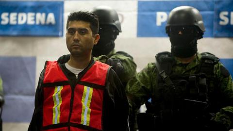 In this file photo from 2012, Mexican soldiers are seen escorting Valencia Salazar after his arrest that year. He is wearing a high-vis vest over a dark hoodie and a white T-shirt. The two soldiers behind him are wearing helmets, dark glasses, and balaclavas. They are armed and are wearing bullet-proof vests. 